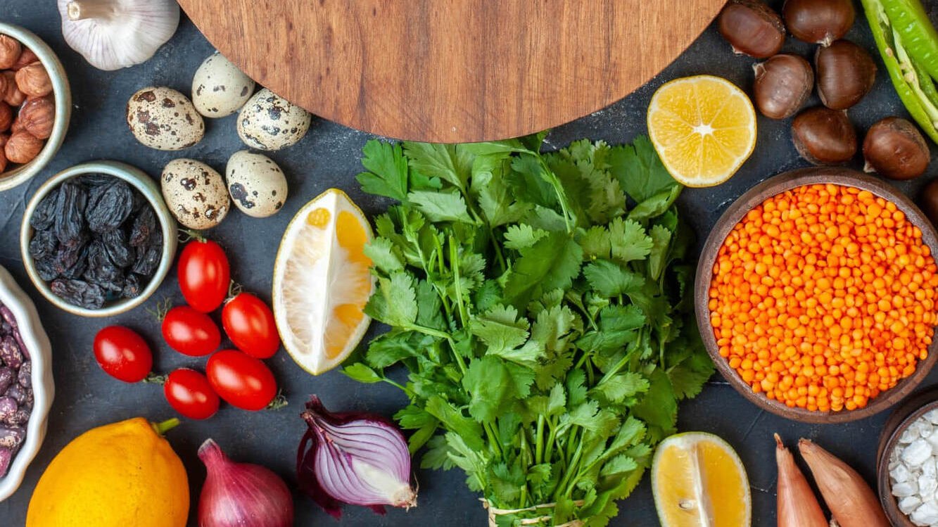 Comida sobre la mesa. Tabla de madera en el centro y al rededor hay medio limón, perejil, frijoles, cebollas, ajo, champiñones, tomate cherry, especias, entre otros, todo listo para empezar a cocinar.